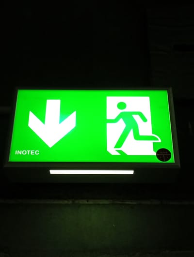 This image, captured in Kabile, Bulgaria, displays a brightly illuminated green emergency exit sign against a dark background. The rectangular sign features two prominent white symbols: a large downward-pointing arrow on the left, and a stylized figure of a person running through an open doorway on the right, indicating an escape route.
Below the arrow, the word "INOTEC" is visible in white capital letters. In the bottom right corner of the sign, superimposed over the running figure symbol, is a small circular sticker with black text. On the sticker, "2R/SSL" is legible, along with the numbers "2" and "G" and "4". Directly below the main sign, a thin, horizontal strip of white light is visible, providing additional illumination to the immediate foreground.
The surrounding environment is predominantly dark, suggesting low ambient lighting, likely indicating nighttime or an unlit indoor space such as a corridor, basement, or parking garage. The sign's strong luminescence sharply contrasts with its dim surroundings, effectively highlighting the emergency escape direction. The scene conveys a functional, safety-oriented environment, with the sign serving as a crucial guide for potential emergencies, directing occupants to descend or exit.