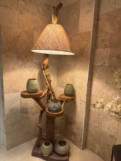 The image depicts an interior scene featuring a unique floor lamp with a burlap-like lampshade and a rustic wooden stand made of branches. The lamp is adorned with several decorative, ornate lanterns of varying sizes, positioned on small wooden shelves attached to the stand. The background is a wall covered in large, light-brown, textured stone tiles. To the right of the lamp, a vase with artificial white flowers is visible. The floor appears to be light-colored marble or polished stone. The scene suggests a decorative corner in a home or a public space, possibly a hotel lobby or a cafe, given the upscale materials and decorative elements. The lighting is warm, indicating artificial illumination from the lamp itself. There are no people present in the image. The overall aesthetic is natural and somewhat bohemian, with a focus on organic materials and intricate decorative details. This scene could be located in Tirat Karmel, Israel, as part of an interior design scheme that incorporates natural elements and artisanal touches.