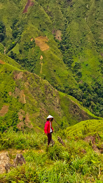 A person wearing a white conical hat and a red jacket stands on a grassy hillside overlooking a valley in Bắc Yên, Vietnam. The surrounding mountains are covered in dense green vegetation and feature terraced fields. The person is positioned in the lower center of the frame, looking towards the right. The environment is natural and mountainous, with a clear sky.