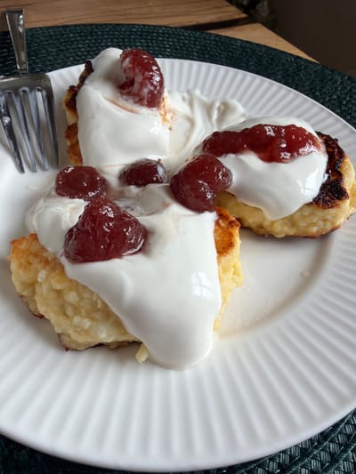 Three syrniki, a type of Russian cheese pancake, are arranged on a white ribbed plate. They are topped with thick white cream and garnished with red berries, likely lingonberries or cranberries. A silver fork rests to the left of the plate. The plate is on a dark green placemat. The background suggests a wooden surface, possibly a table. There are no people visible in the image. The scene appears to be a close-up, overhead shot of a breakfast or dessert. The specific location within Kerro, Russia, cannot be determined from the image.