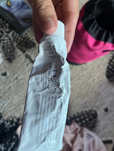 The image shows a close-up of a person's hand holding a tube of Dr. Althea 147 Barrier Cream. The cream tube is white and slightly crinkled, with black and pink text. The text on the tube indicates it is for normal to dry skin types and lists ingredients such as avocado fruit extract, brassica oleracea acephala leaf extract, and various ceramides and hyaluronic acids. The background is blurred but appears to be an indoor setting with a patterned surface, possibly a carpet or textile. A bright pink and black object, possibly a bag or piece of clothing, is visible in the upper left corner. The lighting suggests it is daytime, but no specific time or weather conditions can be determined. The focus is on the product and the hand holding it, suggesting the image is likely for product review or promotion. No faces are visible, and the overall mood is neutral and informative.