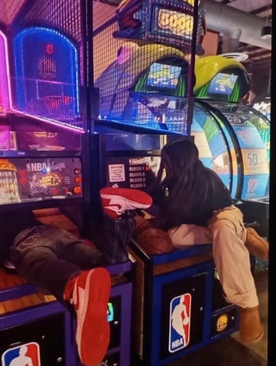 The scene is an indoor arcade setting in Beklar, Uzbekistan, characterized by dim ambient lighting and prominent neon illumination from multiple arcade machines. Two individuals are present. On the left, the lower torso and legs of a person are visible; they are wearing dark jeans and red and white athletic shoes, positioned horizontally across the base of an NBA-themed basketball arcade game. On the right, a second individual is seated on the console of an adjacent NBA-themed basketball arcade game. This person wears a dark long-sleeved top, light-colored trousers, and tan boots, with long dark hair partially obscuring their face as they look towards the machine. A brown basketball rests on the arcade console next to the seated individual. Another red and white athletic shoe is perched on a black vertical partition between the two individuals' arcade units. The arcade machines feature NBA logos, digital displays showing game information, and protective netting. Exposed ductwork is visible in the industrial-style ceiling in the background.