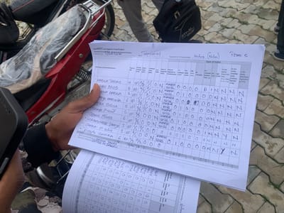 The image depicts a close-up, overhead view of a handwritten form titled "Sanitation and Hygiene Fund Project - Household Committee - Monitoring and Evaluation Checklist." The form is being held by a person's hand, partially obscuring the lower portion of the sheet. The form is filled out with various entries related to households, including names, population counts, and details about toilet facilities. The upper section of the form shows fields for "Name of LGA," "Ward," and "Name of CLTS Community." In the visible portion, "BALZWA" is written under LGA and "TAPSHIN" under Ward. The CLTS Community is written as "Nature (Naturi)" and the team as "Team S." The main body of the form is a table with columns for "S/No," "Name of Household/Institution," "Popn of HH," "Gender Adult Popn (M/F)," "Children Popn (M/F)," "# of PWD," "New or Upgrade to Toilet," "Date of Construction," and various types of toilets such as "Pit without cover," "Pit with cover," "VIP (with vent)," "PF-Ceramic," "PF-cement/ PF-broken Tiles, etc," "WC," "Is the toilet Twin Pit? (Y/N)," "Is the toilet SHARED with another family? (Y/N)," and "Is the toilet overflow/ng/leaking (Y/N)." Several entries are visible in the table, with the first entry being "HABILA TAKAS" with a population of 12 and details about the toilet type. Subsequent entries include "AHILI DAVID," "SHUA HABILA," "ADIYA HARUNA," "APAS BULUS," "ISHAYA PIWULT," "GIDEON PIHIIR," "LOKEMBHIR ISHAYA," "ZAKKA P. IWULT," and "JOSEPH ZAKKA." Most of the toilet type columns are filled with "N" or "0," indicating no or specific toilet types. The lower part of the image shows another section of the same form, or possibly a second sheet, with more tabular data, though less clear. The background of the image reveals a red and black motorcycle, suggesting the scene is outdoors, possibly in a public space or a residential area. The ground appears to be paved with interlocking bricks. The presence of the motorcycle and the overall lighting suggest an outdoor environment. The person holding the form is wearing a patterned long-sleeved shirt. The context of the form and the handwritten entries suggest a community survey or data collection related to sanitation and hygiene in Nigeria. While the image does not explicitly state the city of Wai A, Nigeria, the presence of "BALZWA" as the LGA name strongly suggests the location is within the Balan Local Government Area, which is a part of the Gombe State in northeastern
