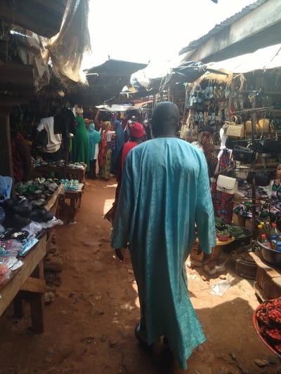 The image shows a busy market street in Lokoja, Nigeria, located at latitude 7.404 and longitude 7.632. It is a narrow, dusty pathway lined with vendors selling various goods, including sandals and handbags.  Several people are walking along the street, many of them wearing traditional clothing. One individual, a man, is walking towards the camera, dressed in a long, blue robe. In the background, other people are engaged in shopping and conversation. The market is partially covered by canopies and shelters. The vendors' displays include an assortment of goods, along with baskets and crates.  The scene is bustling and active.