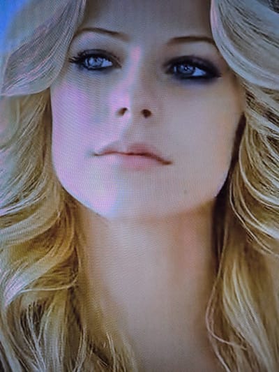 The media file is a close-up, rotated image of a person's face, likely a woman given the soft features and blonde hair. The subject's face is turned to the side, with one eye prominently visible and the other partially obscured by hair. The eye is blue, with visible eyelashes and a reflection in the pupil. The skin is fair, with a slight pinkish hue. A hint of the nose and mouth are also visible. The hair is long and wavy, a light blonde color, and frames the face. The lighting appears soft, suggesting an indoor setting or possibly a cloudy day, though no specific time of day can be determined. There are no discernible objects, text, or environmental details that would provide further clues about the setting or activity. The image captures a moment of stillness, and the expression is neutral or perhaps introspective.