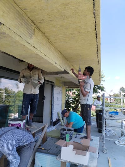 This media file depicts a construction scene outdoors in Dumbrăveni, Romania, under a bright, clear sky, suggesting it is a sunny day. The primary subjects are four men engaged in construction work, seemingly insulating or cladding the underside of an overhang or balcony.

One man, wearing a light-colored shirt and dark shorts, is standing on a makeshift platform and holding a drill, actively working overhead. Another man, in a white shirt and dark pants, stands slightly further back on scaffolding, also appearing to be involved in the construction. In the foreground, a man in a blue t-shirt and dark shorts is using a trowel to apply material, possibly plaster or cement, to the building. He is leaning over a bucket and a work surface. A fourth individual, wearing a plaid shirt and grey pants, is bent over in the foreground, out of focus, with his back to the camera, suggesting he is also contributing to the task.

The setting is an exterior of a building under construction, characterized by exposed insulation material (appears to be yellow mineral wool), white brickwork, and scaffolding. Large windows are visible on the building. Construction materials like buckets, boxes, tools (including a trowel and hammer), and insulation boards are scattered around the work area.

The activity is focused on the application of materials to the building's exterior, likely for insulation or finishing. The workers appear focused on their tasks, with no strong emotional cues visible. The time of day appears to be midday due to the bright sunlight and clear sky. Some visible text includes "RAFT" on a bucket and "HILTI" on the drill.
