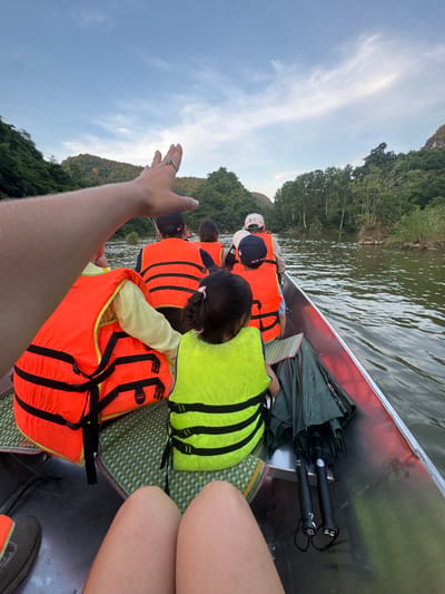 The image shows a group of people on a boat in a river, likely in the city of Yên Sơn Ward, Vietnam, given the vegetation and topography. The boat is a long, narrow vessel, possibly aluminum, with a red interior trim. A person in the foreground is partially visible, with their hand raised towards the sky, wearing a ring with a blue stone. Several other individuals are seated in the boat, all wearing life vests. Some are wearing orange life vests, while others, including two children, are wearing bright yellow-green life vests. The children are seated towards the front of the boat, one with her back to the camera, her hair in a ponytail. In the background, lush green trees and hills line the riverbanks. The sky is overcast with some patches of blue and wispy clouds. Two closed umbrellas are secured in a holder on the side of the boat. The water appears calm with gentle ripples. The overall scene suggests a leisurely boat tour or transportation on a river.