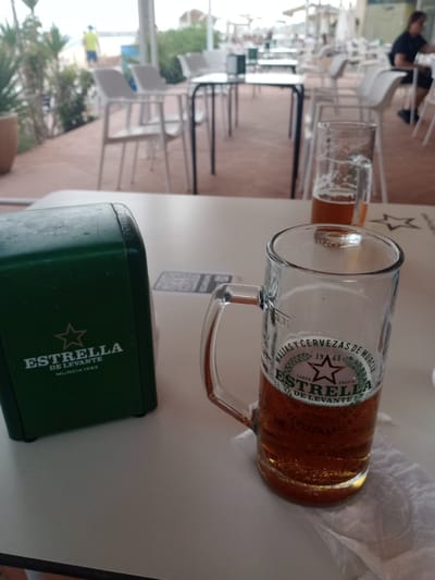 A close-up shot shows a table at an outdoor seating area of a restaurant or bar, possibly near the beach in Águilas, Spain. The table is white and smooth. In the foreground, a dark green rectangular napkin dispenser with a white star logo and the text "ESTRELLA DE LEVANTE MURCIA 1963" is on the left. To the right of the dispenser is a glass beer mug filled with amber beer and a frothy head. The mug has "ESTRELLA DE LEVANTE" printed on it. Behind the mug, a partial view of another glass of beer is visible, as well as a QR code printed on the table. In the background, there are white chairs and tables, and the blurry figures of people seated at them. Beyond that, a beach and the ocean are visible under an overcast sky, with some buildings in the distance.