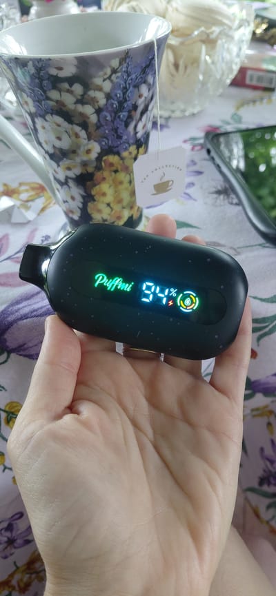The image shows a close-up of a hand holding a black electronic device with a digital display. The display shows the word "Puffmi" in green, followed by "94%" in white digits. To the right of the percentage, there are icons that appear to represent a battery (a red lightning bolt) and liquid level (a green droplet within a circle). The background is blurred, but it appears to be a table covered with a floral-patterned cloth. Behind the hand and device, a white mug with a floral design and a tea bag tag are visible, along with a glass bowl containing what looks like dried flowers or potpourri. A corner of a smartphone is visible on the right side of the image. The image does not contain any information to suggest the location is Tashkazgan, Russia.