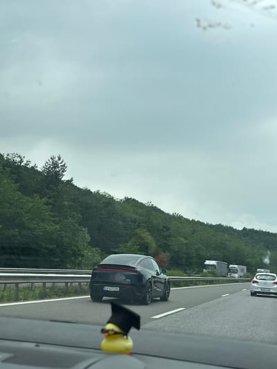 The image depicts a highway scene on an overcast day, observed from inside a vehicle, with a yellow rubber duck figurine wearing a black graduation cap and glasses on the dashboard in the immediate foreground. Ahead, a dark-colored Porsche Taycan, identifiable by its rear design and full-width taillight strip, travels in the left lane. Its Bulgarian license plate reads "EA 6272 AE." Further down the highway in the right lane, a white hatchback, likely an Opel Astra, is visible, with a partially obscured Bulgarian license plate starting with "CA." Two white semi-trailer trucks are seen further ahead in the right lane. The highway features white lane markings and a metallic guardrail on the left. Both sides of the highway are flanked by densely forested hills. Given the "EA" license plate prefix indicating the Blagoevgrad region in Bulgaria, the scene is located on a highway near Gorno Varshilo, Blagoevgrad Province, Bulgaria.