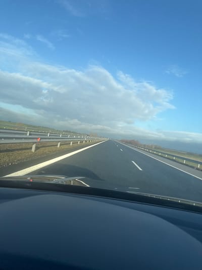 An overhead view from inside a moving vehicle captures a multi-lane highway near Воденичане, Bulgaria, under a partially cloudy blue sky. The dark asphalt road features a solid white line on the right shoulder and a dashed white line marking the inner lane. A continuous metal guardrail with red triangular reflectors runs along the right side of the carriageway, separating it from a grassy verge and a gentle embankment. A similar guardrail is visible on the left, indicating a divided highway. Beyond the right guardrail, a landscape of light brown grass and distant deciduous trees with reddish-brown foliage is apparent. The horizon on the right shows darker, heavier cloud formations, while the upper sky is bright blue with scattered white cumulus clouds. No other vehicles or individuals are present on the visible stretch of the highway. The lower foreground shows the reflection on the dark dashboard or hood of the vehicle from which the image was captured.