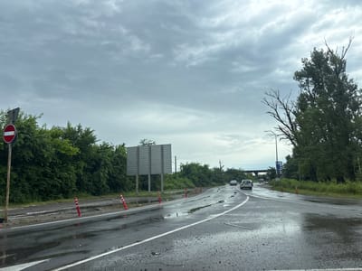 The image depicts a wet, overcast day on a two-lane road with a curve to the right. Orange and white traffic cones are placed along the side of the road, and a large, blank billboard is visible on the left. A red and white "no entry" sign is also present on the left side of the road. Several cars are driving on the road, and puddles of water are on the asphalt, indicating recent rainfall. Lush green trees and vegetation line the sides of the road, and a pedestrian overpass is visible in the distance. The scene is consistent with a road leading into or out of the city of Samtredia, Georgia.