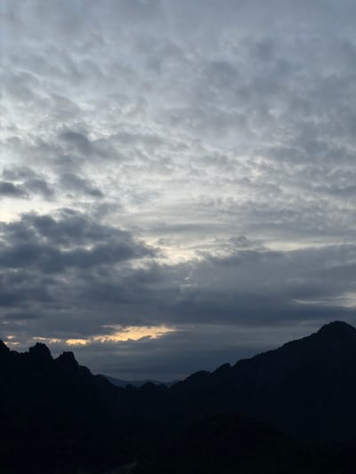 The image depicts a mountainous landscape under a cloudy sky, likely taken at dusk or dawn. The foreground is dominated by the dark silhouettes of rugged mountains, with sharp peaks and dense foliage visible at the base. In the mid-ground, several layers of mountains recede into the distance, becoming progressively fainter and more muted in color. The sky is filled with a textured layer of grey and white clouds, with a band of soft, warm light breaking through in the horizon, suggesting the presence of the sun just below the visible horizon. The scene evokes a sense of natural grandeur and tranquility. The location in Ban Phon Sai, Laos, is known for its dramatic karst topography and misty mountain views, consistent with the visual elements in the image.