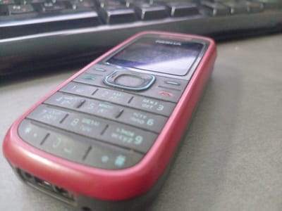 An extreme close-up, high-angle shot shows a red Nokia feature phone placed on a gray desk next to a dark keyboard. The phone is angled with its keypad facing the viewer, and the screen is to the upper right. The keypad is black with white and blue lettering. The numbers 1 through 9, *, 0, and # are visible, along with corresponding letters. The phone has a round, blue-accented navigation button in the center. The keyboard is out of focus and appears to be a standard computer keyboard with dark keys. The background is a neutral gray desk surface. No people or actions are depicted. The image is not specific to Mamonovo, Russia.
