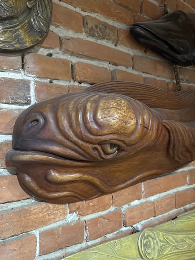 The image shows a close-up of a wooden fish sculpture mounted on a brick wall. The fish sculpture is brown and has visible grain and texture, with carved details suggesting scales and fins. Its eye is a dark circular recess, and its mouth is slightly open. To the upper left, a portion of a gold-colored, ornate decorative object is visible, possibly a mask or a relief. To the upper right, a dark, smooth sculpture of a fish head with an open mouth is also attached to the wall, with a chain visible. The brick wall is made of reddish-brown bricks with white mortar. A portion of a yellow, carved decorative element is visible at the bottom of the frame, featuring floral motifs. The overall impression is of decorative objects displayed indoors, possibly in a rustic or historically themed setting. No specific location in Kolomyia, Ukraine, can be identified from the image itself.