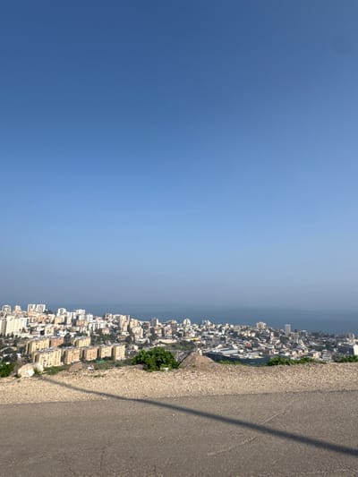 The image presents a panoramic, high-angle view overlooking the city of Tiberias, Israel, situated on the western shore of the Sea of Galilee. The scene is captured under a clear, unclouded blue sky, which dominates approximately the top two-thirds of the frame. A very small, dark, unidentifiable speck is visible in the upper left quadrant of the sky.
In the immediate foreground, a section of dark grey asphalt pavement occupies the bottom of the frame, exhibiting minor cracks and light-colored aggregate. A distinct, elongated dark shadow traverses this pavement diagonally from the lower right towards the middle-left. Beyond the pavement, an unpaved embankment of earth and gravel slopes downward, dotted with scattered rocks and small clusters of green shrubbery.
The midground features the urban expanse of Tiberias, characterized by numerous multi-story buildings constructed on undulating terrain that descends towards the water. These structures vary in architectural style and height, with many appearing as residential blocks in shades of beige, white, and light grey.
The background consists of the expansive, blue-grey surface of the Sea of Galilee, which stretches to the horizon line, meeting the sky without discernible distant landmasses. The overall scene is brightly lit by natural daylight, evident from the crisp blue sky and the clear, defined shadows present. No individuals or specific activities are observable within the frame.