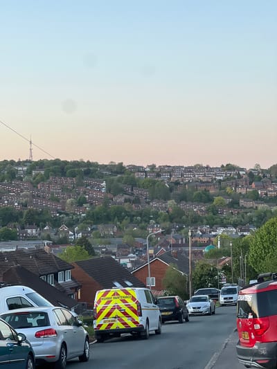 The image, originally oriented sideways, displays a panoramic view of a residential area built on a hillside, characteristic of towns like High Wycombe, United Kingdom. The time of day appears to be either sunrise or sunset, indicated by the soft gradient of the sky, which shifts from a pale blue on the left to a warm, light orange-pink hue on the right.
In the foreground, a paved street is visible, lined with several parked vehicles. Closest to the viewer is the rear of a white van with prominent yellow and red chevron markings and the text "HIGHWAY MAINTENANCE." Its license plate reads "EF71 UXX." Further along the street, a silver car (possibly a Volkswagen Golf variant) and a partially visible dark-colored car are parked. More vehicles are visible further down, including a black car with license plate "LT04 RNH," a white Ford Focus with license plate "WF08 LWC," another white van, and a red commercial van or truck. The red vehicle has partial text, possibly "PLUMBERS," on its side and a license plate "GJ10 EKA."
Beyond the street, the hillside is densely populated with numerous houses, featuring brick roofs and varied architectural styles, largely nestled among abundant green trees. A notable architectural feature, a dome-shaped building, can be seen among the houses on the right side of the hillside. In the far distance, towards the left horizon, a tall, slender mast or tower structure is faintly visible against the twilight sky. Streetlights and overhead utility cables are also scattered throughout the scene. The overall impression is a serene, well-established urban landscape integrated into a natural, hilly environment.