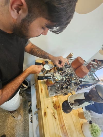 A man with dark hair and a beard stands by a coffee bar, wearing a black shirt and white shorts. He's using a professional espresso machine to froth milk in a small pitcher. The machine sits on a wooden countertop alongside a coffee grinder and a plant. He has a tattoo on his right forearm. His outfit is completed by his Adidas sneakers.
