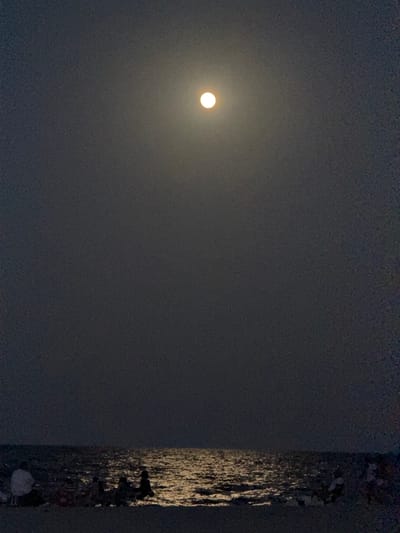 A full moon shines brightly in a dark, hazy sky above a calm ocean at night. The moon is positioned in the upper center of the frame, casting a shimmering path of light across the water. The surface of the water appears to be gently rippling. In the foreground, several people are gathered on the shore, silhouetted against the moonlight. Some individuals are sitting on chairs or blankets, while others appear to be standing. They are facing the water and the moon, seemingly enjoying the nocturnal spectacle. The overall scene conveys a sense of peacefulness and serenity. The location is the city of Hollywood, United States of America.