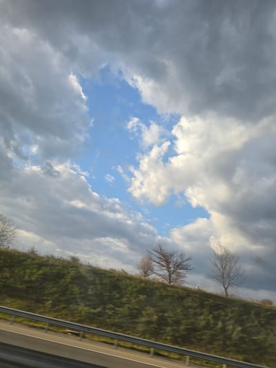 The image displays a landscape captured from a moving vehicle, exhibiting an upward-angled view. The upper half of the frame is dominated by a dynamic sky featuring a mix of dark grey and white cumulus clouds, with a prominent irregular opening of clear blue sky visible in the upper-middle portion, interspersed with smaller white clouds.
The lower half of the image depicts a sloping green hillside covered with sparse, short vegetation, suggesting an early spring or late autumn season. Several bare deciduous trees are positioned on the hillside; at least three distinct trees are visible, one partially cropped on the far left, another in the center-right, and a third on the far right. No foliage is discernible on these trees. A faint, dark horizontal line, possibly a fence or a wire, is visible along the upper edge of the hillside.
In the foreground, a multi-lane asphalt road or highway is partially visible on the bottom left. A silver-colored metallic guardrail runs horizontally across the lower part of the image, separating the road from the hillside. The immediate foreground below the guardrail is dark, likely representing the vehicle's interior or the obscured lower part of the road. No individuals, specific actions, or interactions are observable.
The scene is described as being in Yazdach, Bulgaria. The depicted landscape is consistent with rural or exurban environments found in parts of southeastern Europe. However, "Yazdach" is not a recognized major city or town in Bulgaria, and no specific geographical markers identifying this precise location are visible within the image.