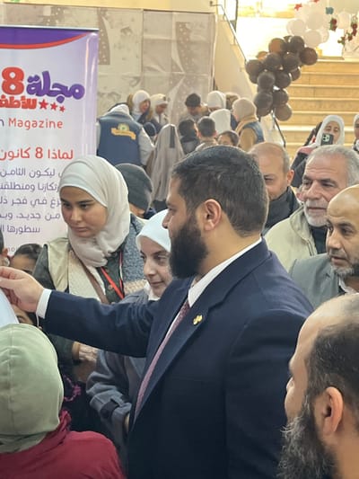A densely populated indoor gathering is depicted in Kafr Batna, Syria. In the foreground, a male subject with a dark beard and dark hair, wearing a dark blue suit jacket, a white shirt, and a burgundy tie with a gold lapel pin, extends his right arm forward, appearing to interact with an individual partially obscured below the frame. To his left, a female subject wearing a light beige hijab and a light-colored vest over a dark garment looks downward and to her left. Behind her, another female subject with a white hijab is observed smiling.
The midground and background are populated by numerous individuals, including adults and children, many wearing head coverings (hijabs) and various winter clothing items. On the far left, a large banner is partially visible, displaying purple and red text in Arabic and English, including "Magazine 8 for Children" and "Why January 8." In the upper right background, a light-colored staircase is present, adorned with clusters of dark brown and white balloons, and star-shaped decorations. The overall environment suggests a public event or celebration within a brightly lit indoor space.