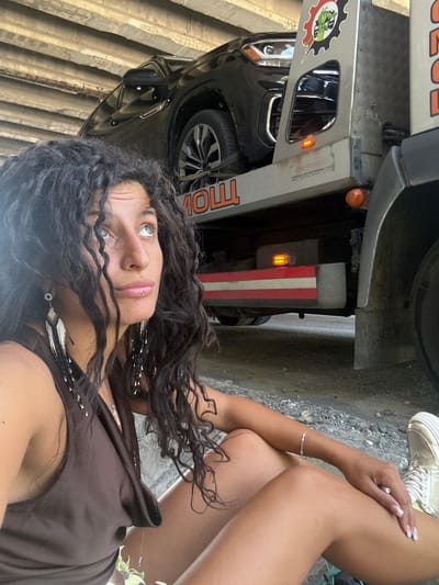The image captures a woman with long, dark, curly hair sitting on the ground in what appears to be an urban or semi-urban environment. She is looking upwards and to her right. She is wearing a brown sleeveless dress and large, beaded earrings. Her right arm is resting on her left leg. To her right, partially visible, is a tow truck. A black SUV is loaded onto the bed of the tow truck. The tow truck has "MOLL" written on its side in orange lettering. The scene is situated under an elevated concrete structure, possibly a bridge or overpass. The ground is rough and appears to be a mix of dirt and gravel. The overall lighting suggests it is daytime. The specific location of Rajana, Bulgaria is not determinable from the visual information within the image itself.