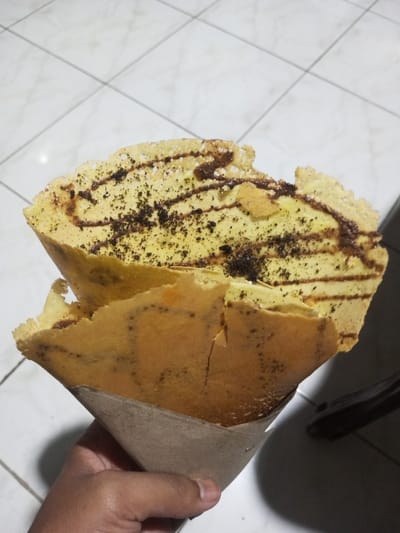 The specific city of Pandaria, India, is not discernable from the visual information provided. The scene depicts a close-up of a human hand holding a dessert resembling a folded, thick pancake or crepe. The dessert has a light brown exterior and a lighter, yellowish interior. Visible toppings on the interior include irregular drizzles of dark brown chocolate syrup, scattered dark granular particles (consistent with crushed cookies or chocolate pieces), and sparse white powder, possibly powdered sugar. The top edge of the dessert appears somewhat irregular with minor tearing. The lower portion of the dessert is encased in a plain, light brown paper wrapper, held by a hand with medium-dark skin tone. The background consists of a light-colored, possibly white, tiled floor with dark grout lines, indicating an indoor or covered outdoor setting. No other subjects or interactions are present.