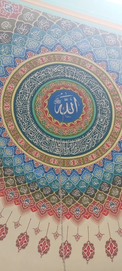 An interior view of a mosque ceiling in Kumbang, Indonesia, displays a large, elaborate circular artwork. The central element is a dark blue disc featuring the Arabic word "Allah" rendered in white calligraphy. This disc is encircled by several concentric rings, each containing intricate patterns and white Arabic calligraphy on backgrounds of teal, red, light green, and yellow-beige. These rings are further surrounded by scalloped, layered patterns in shades of blue, green, brown, and red, each segment containing a stylized Arabic script. The outermost visible portion of the artwork transitions into simpler, hanging decorative motifs in red and beige on a light wall. A thin silver-colored metal chain extends vertically downwards from the central blue disc, partially obscuring the lower part of the Arabic script within the disc. The upper edge of the image shows a plain, light blue and beige wall or ceiling section.