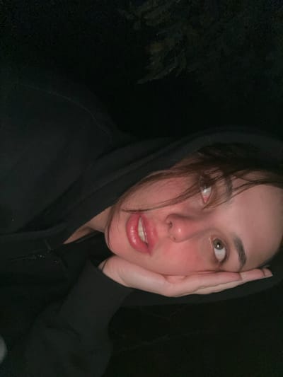 A close-up, angled portrait depicts a single individual, identified as a young female, against a dark, low-light background. The subject's head is tilted right, resting on their left hand which supports the chin. They wear a dark, likely black, hooded garment, with the hood partially framing the face. Facial features include brown hair, light-colored eyes (appearing brown/hazel), and full, slightly parted lips with visible upper front teeth. The skin exhibits some redness on the cheeks and nose. The subject's right eye displays a red-eye effect, indicative of direct flash photography. The gaze is directed upwards and to the right of the frame. The environment, though largely obscured by darkness, shows faint outlines of tree branches or foliage in the upper left quadrant, suggesting an outdoor setting. The scene is illuminated by a direct, bright light source. The image is described as originating from the city of Dibrova, Ukraine.