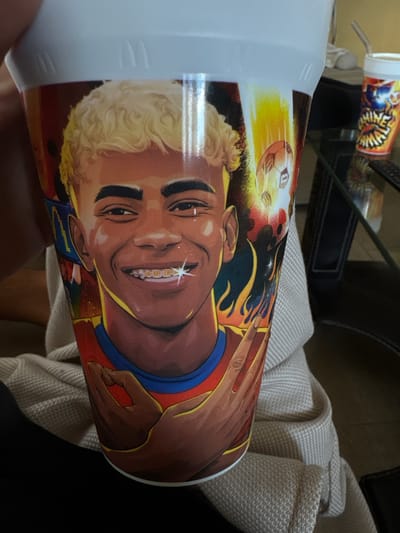 The image shows a white plastic cup with a vibrant graphic wrapping around it. The graphic features an illustration of a young person with curly blonde hair and dark skin, wearing a red and blue outfit. They have a wide smile, revealing braces that are sparkling. Their left hand is raised, with fingers forming a circle, and a starburst is visible near their mouth. The background of the illustration shows a soccer ball in motion against a fiery, yellowish-orange backdrop, suggesting a theme related to sports or energy.
To the left of the cup, a small portion of another cylindrical object with a similar colorful graphic is visible. This object also appears to be a container, possibly another cup or a small bottle.
The cup is resting on a textured, light-colored fabric, which appears to be a blanket or a towel. The background is dark, with some hints of furniture or other objects, possibly in a room. The lighting seems to be indoor, as indicated by the general tone and the reflection on the cup's surface. The overall mood of the illustration on the cup is energetic and joyful. No discernible text is visible on the main cup, but the smaller object in the background might have some lettering on its graphic.
