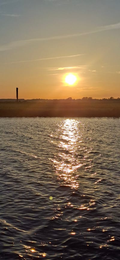 The image depicts a tranquil waterscape at sunset or sunrise. The sun, a bright orb radiating warm yellow and orange light, hangs low in a hazy sky. Its reflection shimmers and dances across the rippling surface of the water, creating a dazzling spectacle of light. The water itself appears dark, with the light catching the crests of small waves, giving it a textured, almost sparkling appearance.
Along the left side of the image, a silhouetted shoreline is visible, softened by the atmospheric light. A dark, rectangular structure, possibly a small pier or marker, juts out from the shore into the water. The landmass is lined with a dense band of trees, their forms indistinct against the glowing sky. The overall mood is one of peacefulness and natural beauty, amplified by the soft hues of the sky and the dynamic play of light on the water. The time of day is clearly indicated by the low position of the sun and the warm, diffused light. The location in Pärnu, Estonia, suggests a coastal or lakeside setting. There is no visible text in the image.