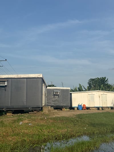 Igwuruta, Nigeria. The image shows a low-angle, side view of a row of prefabricated buildings, likely office or storage units, next to a vegetated area bordering a body of water. Two dark grey, rectangular units are prominent in the foreground. One has a white stripe near the roof and a window-type air conditioning unit installed. Behind these, a light-colored building with closed windows is visible. Next to the light-colored building are two overturned plastic chairs, one blue and one orange. The landscape consists of tall grasses and vegetation, with a water body visible on the right side. A blue sky with light cloud cover is above the buildings. In the background, dense tropical foliage suggests banana trees. Power lines are visible on the left side. The overall impression is of a utility area, possibly a construction site or temporary office space.
