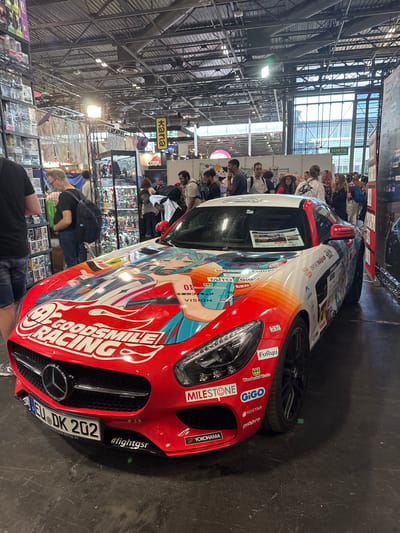 The image depicts a red Mercedes-Benz AMG GT sports car with an anime-inspired livery at a convention in Villepinte, France. The car is prominently displayed and covered in graphics featuring anime characters and various sponsor logos, including "GOODSMILE RACING," "MILESTONE," "FuRyu," "GiGO," and "YOKOHAMA." A license plate is visible on the front bumper with the text "EU DK 202." The scene appears to be indoors, likely within a large exhibition hall, indicated by the exposed ceiling structures, lighting rigs, and various booths and displays in the background. Several people are present, milling around the car and the surrounding exhibits, suggesting a public event or trade show. The environment suggests a bustling atmosphere typical of a convention or exhibition.