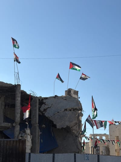 In Jabalia, Palestinian Territories, a building has been heavily damaged and shows signs of structural collapse. The scene is dominated by the clear blue sky and the rubble of what appears to be a destroyed structure. Numerous flags are prominently displayed, attached to poles and strung across the damaged remnants. Among these flags are the Palestinian flag, identifiable by its horizontal stripes of black, white, and green, and a red triangle at the hoist. Other flags visible include what appear to be the flags of Egypt and potentially other Arab nations. Some flags are tattered, fluttering in the wind against the backdrop of the devastated environment. The damage to the building is extensive, with exposed rebar, crumbled concrete, and debris scattered throughout. The overall impression is one of destruction and a defiant display of national symbols amidst ruin.