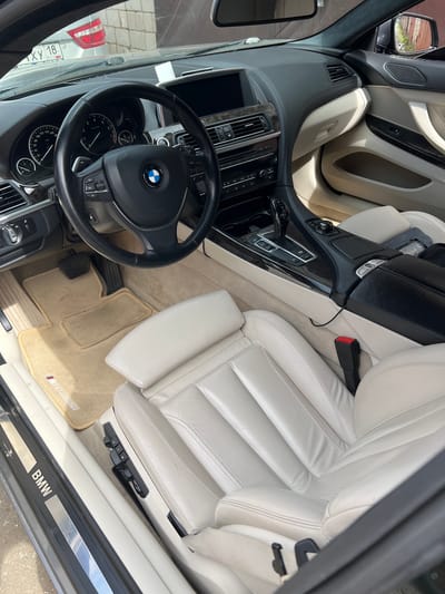 The image shows the interior of a modern white leather BMW, captured from the passenger side, looking towards the driver's side. The car is parked outdoors, with bright sunlight illuminating the interior and casting shadows. A license plate with Russian characters and the text "XY 18" is visible on the windshield of a vehicle in the background, suggesting the location is in Russia. The dashboard is black with wood-grain accents, featuring a central infotainment screen and instrument cluster. The steering wheel is also black leather with the BMW logo in the center. The seats are a light, cream-colored leather. The floor is covered with beige mats, one of which has the "M" logo. The gear shift is visible in the center console. The overall impression is of a well-maintained luxury vehicle.
