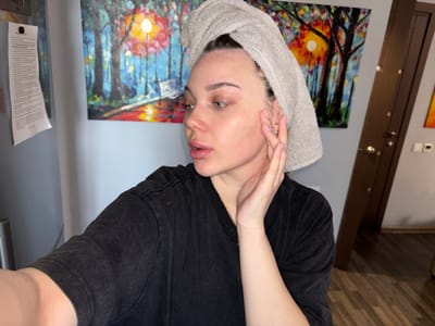 A young woman with a towel wrapped around her head is looking to her left, her right hand touching her face. She is wearing a black t-shirt. Behind her, on the wall, are two abstract paintings, one depicting a colorful forest scene with a bright sun, and another partially visible to the right. A white piece of paper is attached to a surface to the left of the woman. The environment appears to be indoors, possibly a room in a residence. The location is identified as Voronovka, Russia.