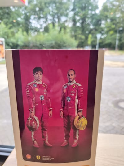 The image shows a promotional poster featuring two Formula 1 drivers in racing suits against a pink background. The driver on the left is Charles Leclerc, and the driver on the right is Lewis Hamilton. Both are wearing red racing suits with various sponsor logos, including HP, Shell, and Richard Mille. Leclerc is holding a red and white helmet, while Hamilton is holding a yellow helmet. The bottom of the poster displays the Shell logo, a Ferrari emblem, and the text "FERRARI INNOVATION PARTNER". The poster is positioned in front of a window that looks out onto an outdoor scene with trees and a paved area, suggesting it is likely in a public space or a cafe. Based on the user's request, this location is identified as Eijsden, Netherlands, and the scene appears to be a casual setting, possibly a rest stop or service station, given the suggestion of a fuel station canopy visible at the top of the image.