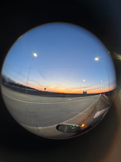 The image shows a fisheye view of a highway at sunset, likely reflected in a spherical object. The sky transitions from blue to orange near the horizon. A row of streetlights lines one side of the highway. A vehicle is visible on the right side of the highway. The image is taken in Kuregovo, Russia.