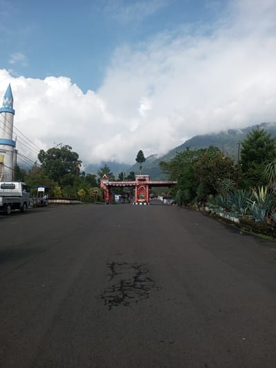 The image depicts a road leading towards a gate in Mya Sit Kaing Ywar Thit, Myanmar. The road is asphalt and shows signs of wear. A light gray utility vehicle is parked on the left side of the road near a blue-and-white tower. The gate is reddish-pink and features the text "SELAMAT DATANG". Beyond the gate, there is a mountain covered in green vegetation and partially obscured by clouds. The sky is a mix of blue and white clouds. On the right side of the road, there are various plants and trees.