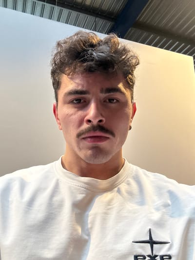 An adult male with fair skin, dark curly hair, a dark mustache, and dark eyebrows is depicted in an indoor setting in Chantepie, France. He wears a light-colored, possibly off-white, long-sleeved crew-neck shirt or sweatshirt featuring a dark embroidered logo on the lower left chest, comprising a cross-like symbol above the letters "PXP." A small silver hoop earring is visible in his left ear. The subject maintains a neutral expression, looking directly into the camera. The background consists of a plain, light-colored wall or partition, with a corrugated metal ceiling and structural elements visible above, suggesting a commercial or industrial building interior. The lighting appears even, with a warmer tone on the right side of the background.