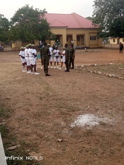 The image captures an outdoor scene at what appears to be a training ground or campus in Ayere, Nigeria, likely in the late afternoon or early morning due to the soft, diffused lighting.

In the foreground and midground, a group of people are gathered on a dry, earthy patch of ground with sparse grass. Several individuals are dressed in military camouflage uniforms, including hats, and appear to be instructors or officers. They are interacting with a larger group of trainees or cadets, who are uniformly dressed in white t-shirts, white shorts or trousers, and green caps. Some of the trainees have small pouches or fanny packs around their waists. They are standing attentively, suggesting an instructional or briefing session is taking place.

In the background, a single-story building with a reddish-orange hip roof and light yellow/tan walls stands prominently. Several windows and doors are visible on its facade. Large green trees flank the building, providing shade. The ground around the group is predominantly dry earth, with some patches of light green vegetation and low stone or concrete borders forming partial circles. A small pile of white, granular material (possibly lime or chalk) is visible on the ground in the lower right foreground. In the far background, another individual in dark clothing can be seen standing.

The overall setting suggests a structured, institutional environment, likely a military or paramilitary training facility. The visible text "Infinix NOTE 8i" is present as a watermark in the bottom left corner of the image.