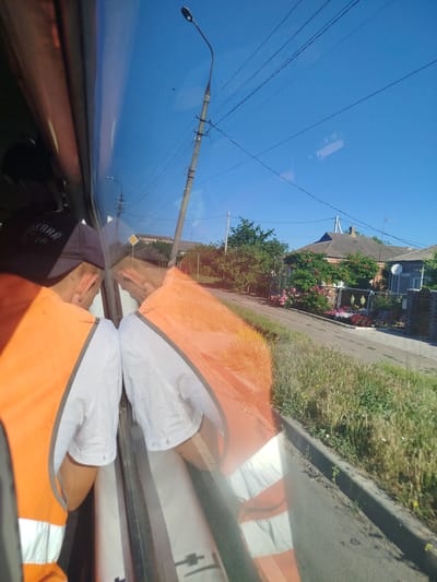The image, taken from inside a vehicle, shows a reflection on the window. In the foreground, two individuals wearing orange safety vests over white shirts are visible through the reflection. One individual has a dark baseball cap on, and the other's hair is lighter. The reflection also shows a street, a sidewalk with overgrown grass and weeds, a road, and residential buildings. The buildings appear to be single-story houses with tiled roofs, set back from the street behind fences. There are trees and flowering bushes in front of the houses. A utility pole with electrical wires and a street lamp are visible in the sky. The sky is clear and blue, suggesting it is daytime. The overall scene depicts a rural or semi-rural area, possibly a village or the outskirts of a town, with elements of daily life such as residential housing and infrastructure. The location is specified as Manhush, Ukraine.