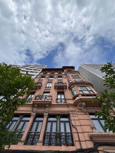 A low-angle, upward-oriented shot presents a building in La Coruña, Spain. It features a facade with an intricate, ornamental design, exhibiting a blend of architectural styles including Beaux-Arts and Art Nouveau, with arched windows and ironwork balconies. The building is adorned in shades of pink and brown, suggesting a possible restoration or preservation effort. Adjacent to this structure is a modern building with a simple white facade, possibly a contrast in styles. The sky, with scattered clouds, fills the upper part of the frame. Green foliage from nearby trees is also visible. The image gives an impression of architectural preservation and juxtaposition within the city.