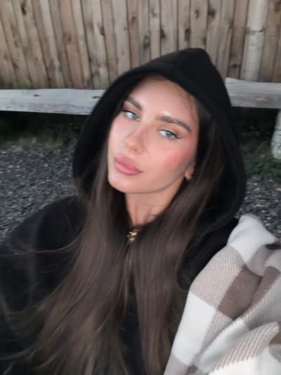 A young woman with long, dark brown hair, wearing a black hooded sweatshirt and a checkered blanket, is pictured in an outdoor setting. She is looking towards the camera, with her face slightly angled. Behind her, there is a wooden fence and a rough, gravel-like ground. The lighting suggests it might be dusk or an overcast day. The location within the city of Манский, Russia is not discernible from the image itself.
