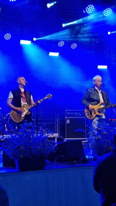 In Priekule, Latvia, two men are performing on a stage illuminated by bright blue lights. The man on the left, wearing a dark vest over a white t-shirt with a graphic, is playing a golden electric guitar and singing into a microphone. The man on the right, dressed in a denim jacket over a white shirt and jeans, is playing a bass guitar. Behind them, stage equipment, including speaker cabinets and lighting rigs, is visible. The stage is decorated with plants in dark pots, adding a touch of nature to the scene. The overall atmosphere appears to be a live music performance.