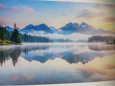 The image depicts a serene mountain landscape at dawn or dusk. Majestic, mist-shrouded mountains rise in the background, their peaks catching the soft, warm hues of the rising or setting sun. In the foreground, a calm lake perfectly mirrors the mountains and the sky, creating a symmetrical and tranquil scene. Dense evergreen forests line the shore, with a few trees standing out in the foreground. The sky is a gradient of soft blues, pinks, and yellows, with wispy clouds adding texture. The overall atmosphere is one of peace and natural beauty. There are no people or identifiable structures from the city of Znamensk, Russia, visible in this image.