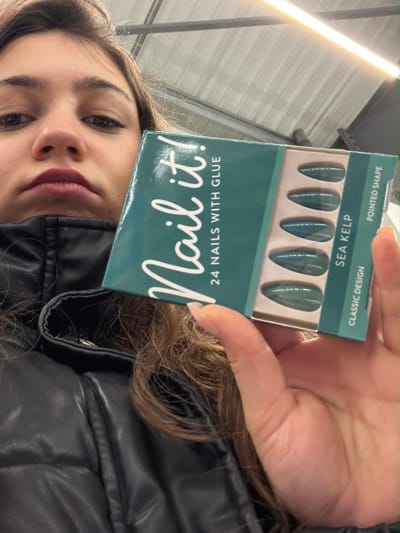 A person holds a box of "Nail it!" brand artificial nails in "Sea Kelp," a set of 24 pointed-shape nails with glue. The box is green with white lettering and features a display of the nails inside. The person is wearing a black leather jacket and has brown hair. A fluorescent light fixture is visible in the background. The person is looking at the camera.