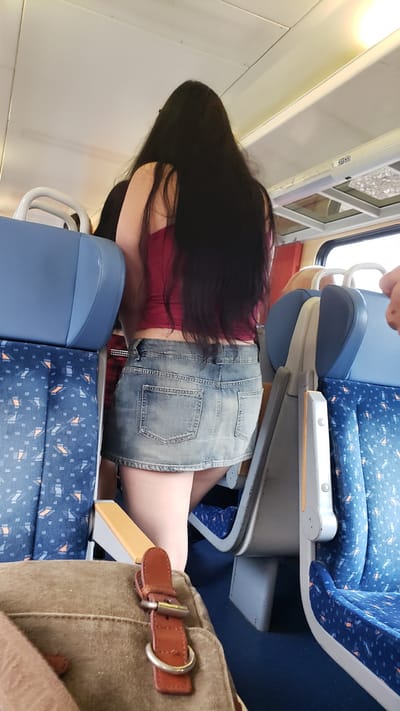 The image depicts the interior of a train carriage, possibly in Liptovský Mikuláš, Slovakia, as evidenced by a sign on the ceiling reading "ST/33". The focus is on the rear view of a person with long, dark hair, wearing a short denim skirt and a crimson tank top. They are standing between blue train seats with a patterned upholstery. In the foreground, a brown canvas bag with a leather strap and buckle is visible. The lighting suggests it is daytime, with natural light entering from the windows. The overall environment is consistent with public transportation.