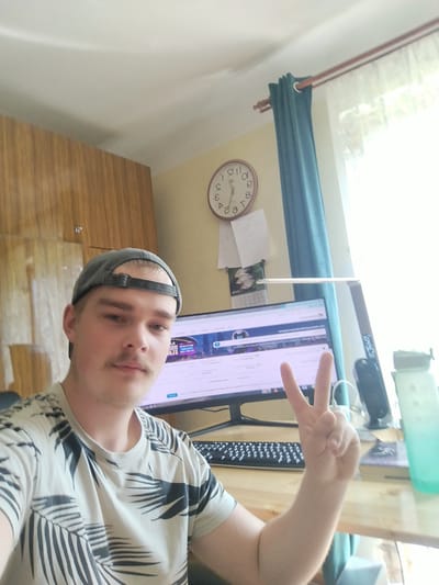 A young Caucasian male wearing a grey baseball cap and a palm-leaf patterned t-shirt is pictured in a selfie taken indoors, presumably in Cēsis, Latvia. He is sitting at a wooden desk in front of a computer monitor. The man is making a peace sign with his right hand. To his left, a portion of a wooden cabinet is visible, and to his right, a window with teal curtains and bright daylight filtering through. A clock on the wall shows approximately 10:07. Other items on the desk include a keyboard, a speaker, a water bottle, and what appears to be a book. The overall setting suggests a home office or study environment.