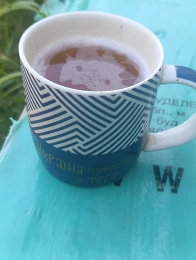 A close-up, overhead shot shows a blue and white patterned ceramic mug filled with a frothy, amber-colored liquid, likely tea or coffee. The mug is placed on a turquoise surface, possibly a table or book. Faint text is visible on the mug and the turquoise surface. The text on the mug appears to be in Ukrainian, with yellow lettering that reads "Україна починається з тебе" (Ukraine begins with you). On the turquoise surface, a large, dark "W" is visible, along with some faint, unreadable text. The background is blurred, with hints of green foliage, suggesting an outdoor setting. The scene is likely located in Ivankovychi, Ukraine, given the text on the mug. The lighting suggests it is daytime. No people are visible in the frame.